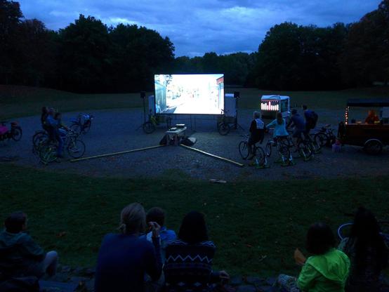 In der Mitte eines Freilufttheaters steht eine Leinwand mit Beamer, auf der ein Film läuft. Auf beiden Seiten daneben sind jeweils 4 Menschen auf Fahrrädern auf Rollen, die Strom erzeugen. Davor sitzen Menschen, die den Film schauen.
