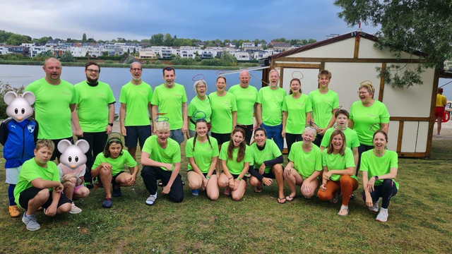 Das Bild zeigt eine Gruppe von etwa 20 Personen, die vor einem See und einer kleinen Hütte stehen. Alle tragen grüne T-Shirts, was auf eine gemeinsame Aktivität oder Gruppenzugehörigkeit hinweist. Vorne sitzen und knien einige Personen, während die meisten im Hintergrund stehen. Einige Personen tragen Kopfbedeckungen wie Hörner und Masken, was auf eine lockere und fröhliche Atmosphäre schließen lässt.