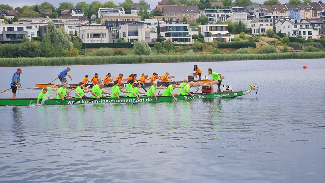 Das Bild zeigt eine Gruppe von Ruderern in einem langen, grünen Drachenboot auf einer ruhigen Wasserfläche. Die Ruderer tragen leuchtend grüne oder orangefarbene Trikots und sitzen in einer Reihe, während sie mit Ruderern synchron arbeiten. Im Boot stehen zwei Personen, die die Ruderer anleiten.