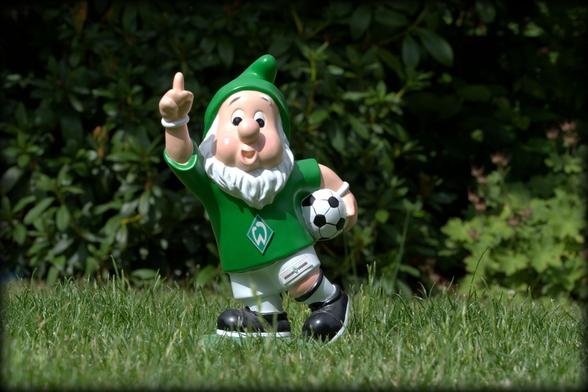 Ein Zwerg mit grüner Zipfelmütze im Werder-Trikot mit einem Fußball unter dem Arm steht auf dem Rase