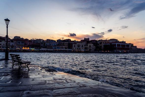 Evening view of Chania’s Old Town waterfront on Crete, with historic buildings and cafés in the distance, and a bench beside a lamp post in the foreground.