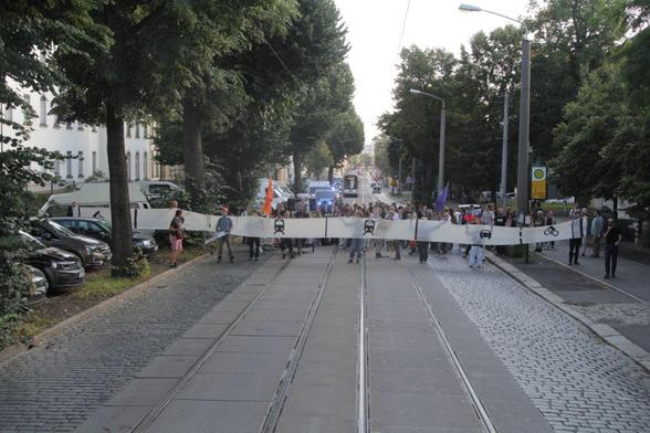 Eine große Gruppe Menschen steht auf der Königsbrücker Straße. Sie halten ein langes Banner, auf dem die Straßenaufteilung der geplanten Straße zu sehen ist. Das Banner ist viel länger als die jetzige Straße.