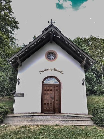 Eine weiße Kapelle mit Kreuz auf dem Dach. Über dem Türbogen der Holztür steht Schriftzug "Nazareth-Kapelle"