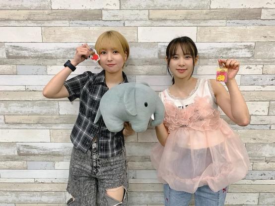 Two young women are standing against a wall with a wooden plank design. The woman on the left, with short blonde hair, is holding a plush elephant and a red keychain. The woman on the right, with long dark hair tied back, is wearing a pink frilly top over a white shirt and holding a pink keychain.