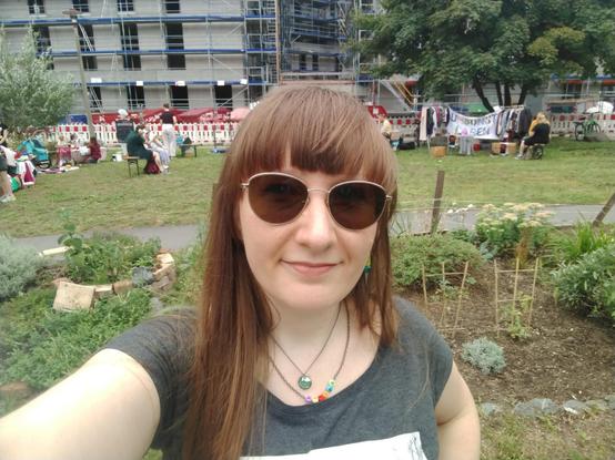 Ich, rotbraune Ponyfrisur und Sonnenbrille, in einem Park mit Ständen im Hintergrund.