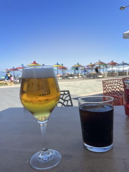 Zwei Getränke auf einem Tisch, im Hintergrund ganz viele Sonnenschirme und Liegen am Strand.
