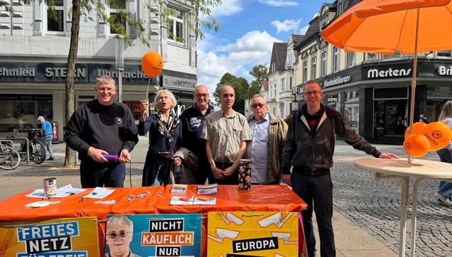 Foto vom Infostand der Piraten Herne: Sechs Leute, zwei davon mit orangen Luftballons stehen hinter einem Tisch mit Aufklebern, Flyern, Plakaten uvam. Rechts steht noch ein oranger Sonnenschirm und ein kleines Tischchen mit orangen Luftballons. Hinter der Piratengruppe sind Geschäfte zu sehen.