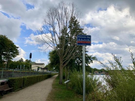 Ein Kiesweg führt auf ein kleines Bahnhofsgebäude zu. Rechts das Ufer des Sees , von Bäumen gesäumt.