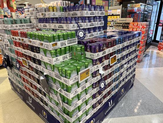 A display of assorted beer cans organized in stacks with various flavors and labels from the brand Unibroue. The cans are grouped by color, showcasing different varieties such as IPA and NEIPA. Several prices are visible on the display, and there are