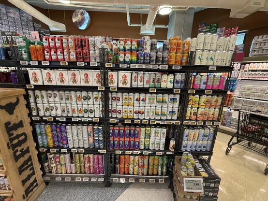 A display of various canned beverages is organized on shelves in a store. Different brands and flavors are visible, showcasing colorful packaging. A shopping cart is partially visible in the foreground.