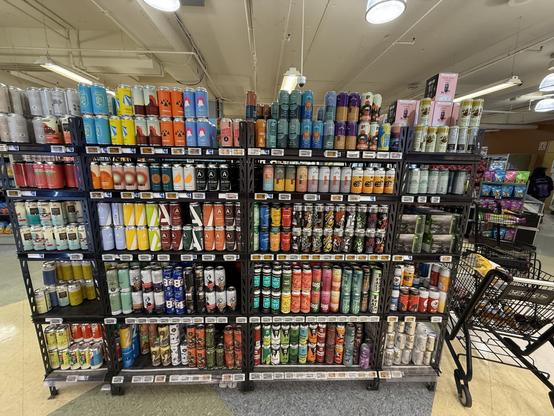 A well-organized display of various canned beverages, including beers and seltzers, on multiple shelves in a store. The cans feature colorful designs and brands, with a shopping cart visible in the foreground.
