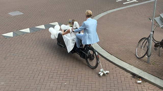 Bride in white dress riding in a bakfiets that is trailing metal cans. One has fallen off in the gutter.