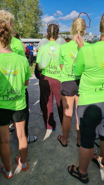 Man sieht mehrere Menschen von hinten mit neongrünen T-Shirts. Auf denen steht Holy Phoenix darüber ist ein weißes Kreuz mit bunten Flügeln zu sehen.