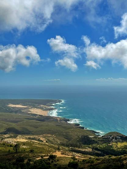 Photo of the ocean shore on the distance. The photo is taken from a high viewpoint. The sky has some scattered clouds but it’s sunny