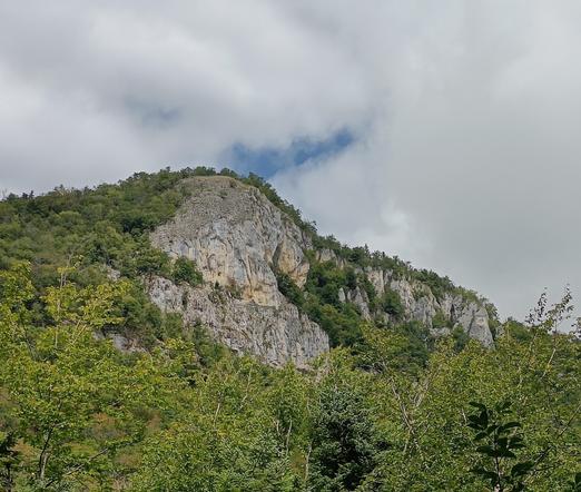 Photographie numérique couleur en format paysage, avec une falaise et un sommet calcaires où se situe le belvédère du Sérémont. La vue est prise en contrebas, avec de la végétation et un ciel bleu couvert de nuages blancs.