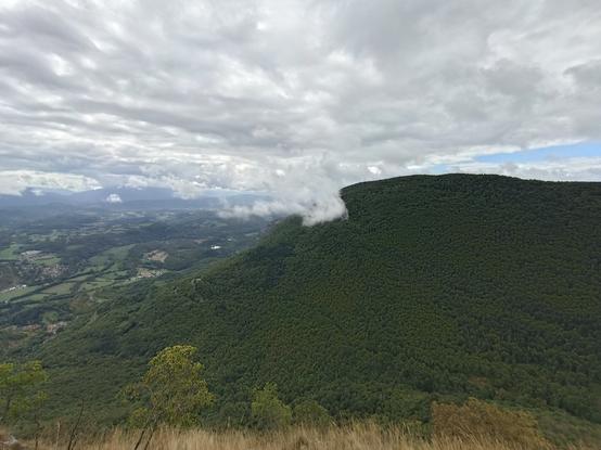 Vue large prise depuis le belvédère, avec la grande montagne de Virieu en face, entièrement boisée, et l'humidité qui s'en échappe et fait grossir un grand nuage gris-blanc. On aperçoit des collines et des prairies en contrebas.