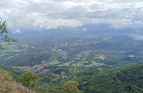 Vue large prise depuis le belvédère, avec en contrebas le bourg de Virieu, des collines et des prairies et au loin, différentes montagnes bleu foncé. Le ciel est chargé de nuages blancs et gris.