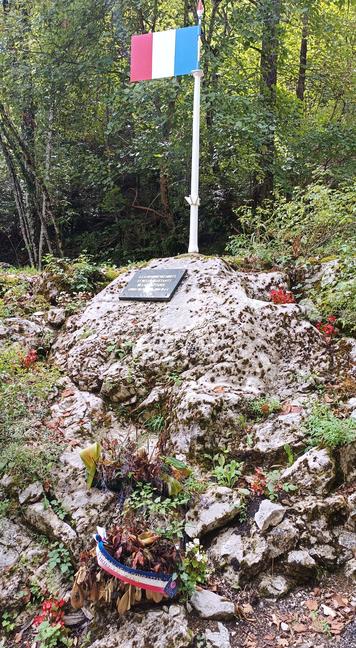 Un monument constitué d'un rocher arrondi, surmonté d'un drapeau français et d'une plaque écrite en doré sur fond noir. Une couronne de fleurs en partie fanées est posée à sa base. Des fleurs rouges sont plantées et il y a des arbres derrière le monument.