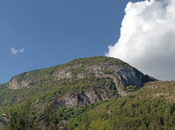 Le sommet d'une montagne calcaire, avec à droite de grands plis convexes de couches de roches. De la végétation et un ciel bleu avec un nuage blanc.