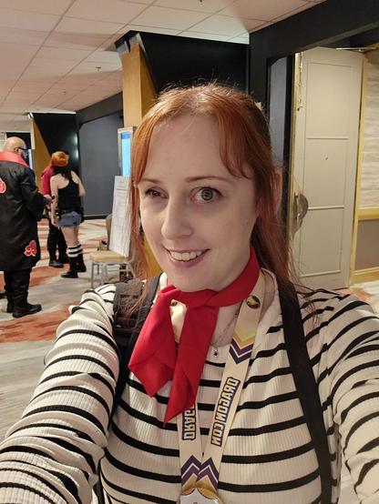 A woman posing for a selfie dressed as Maelle from Expedition 33. She is standing in a crowded hotel lobby