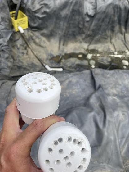 A close-up of me holding to 2 inch pipe caps drilled with many small holes. In the background is the bottom of the pond. The hose from the second pump is visible going down into the bottom of the pond from the yellow crate