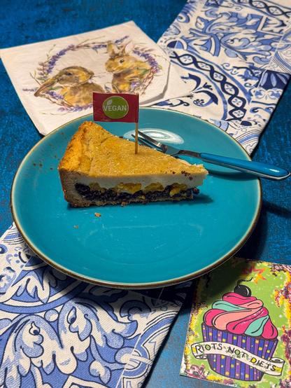Das Bild zeigt ein Stück veganen Kuchen auf einem blauen Teller. Der Kuchen hat eine goldbraune Oberfläche und eine dunkle Mohnschicht Ein kleines "VEGAN" Flaggenstäbchen steckt in dem Stück, um seine veganen Eigenschaften zu kennzeichnen.  Der Teller steht auf einem blauen Tisch mit einem blau-weiß gemusterten Tuch. Im Hintergrund sind ein serviettenartiges Tuch mit einer Zeichnung von Hasen und ein buntes Kartenblatt mit einem Cupcake und der Aufschrift "RIOTS NOT DIETS" zu sehen.