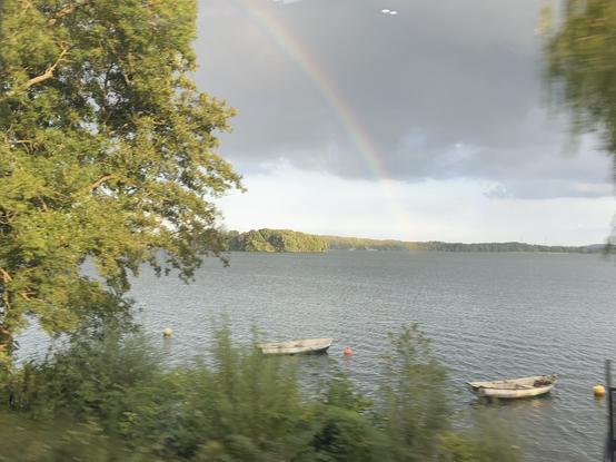 Zwischen gelbgrün scheinenden Bäumen, auf dunkler Wolke über dem See, ist ein halber Regenbogen. Im Wasser liegen einige kleine Boote.