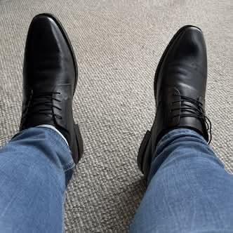 My feet wearing black dress shoes, at the end of my legs with blue jeans, over textured carpet.