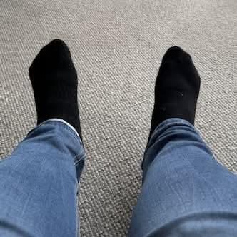 My feet wearing black socks, at the end of my legs with blue jeans, over textured carpet.