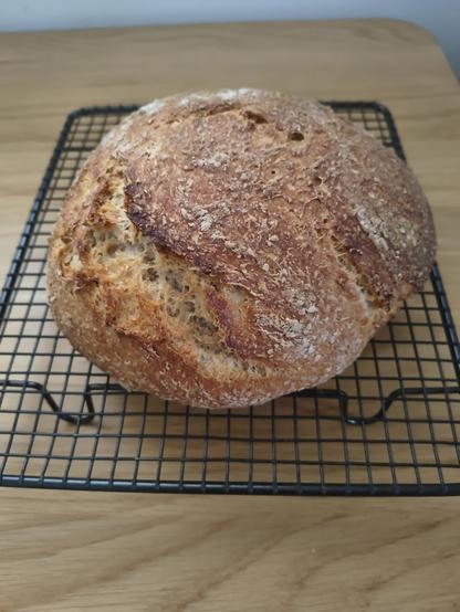 A rounded loaf of bread sitting on a black wire rack, cooling.

The bread is various shades of brown, with a rising related crease on one side which is a lighter shade of brown.