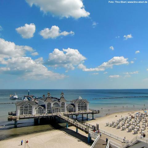 Ein elegantes, weißes Gebäude mit mehreren Türmchen steht auf Pfählen im flachen Wasser und ist über einen langen Holzsteg mit dem Sandstrand verbunden. Rechts davon befinden sich zahlreiche Strandkörbe in Reih und Glied sowie einzelne Strandbesucher. Am Ende des Stegs liegt ein Schiff an einer kleinen Anlegestelle. Das Meer ist ruhig und reicht bis zum Horizont, der Himmel ist blau mit verstreuten weißen Wolken.