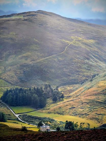 A photograph of a sprawling green and brown hillside with a winding path leading to the summit. A small white house with a patch of trees sits in a valley at the foot of the hill, with a narrow road snaking past it. The sun illuminates a patch of green grass near the house and casts long shadows across the hills. The sky is partially cloudy and a soft light bathes the entire scene.