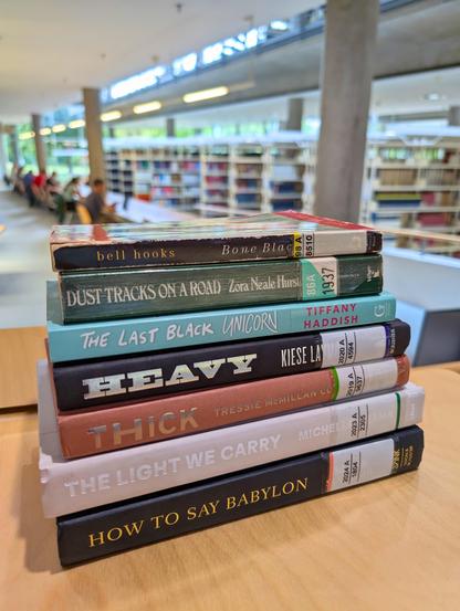 A bookstack of memoirs by Black anglophone writers in the Central Library Building of SUB Göttingen. The room is spacious and bright, with many bookshelves and wooden tables of people working in the background.