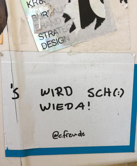 Ein Aufkleber auf einer hellen Oberfläche trägt folgenden Text in Schwarz auf weißem Grund: "'s wird scho wieda! @c.freude". Der Aufkleber ist rechteckig und hat rechts und unten eine blaue Einfassung. Darüber sind Reste eines anderen Aufklebers erkennbar.