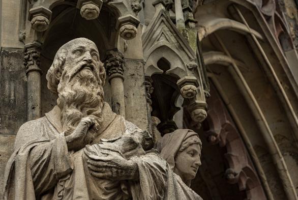 DE: Portrait of the figure of Simeon looking upwards on the exterior of St. Thomas Church in Leipzig. His bearded head gazes into the distance, and he holds the baby Jesus in his left arm. To his left stands the Virgin Mary, looking down at the people standing in front of the Apostle Portal. In the background, you can see the ornate pointed arches of the church entrance.

EN: Portrait of the figure of Simeon looking upwards on the exterior of St. Thomas Church in Leipzig. His bearded head gazes into the distance, and he holds the baby Jesus in his left arm. To his left stands the Virgin Mary, looking down at the people standing in front of the Apostle Portal. In the background, you can see the ornate pointed arches of the church entrance.
