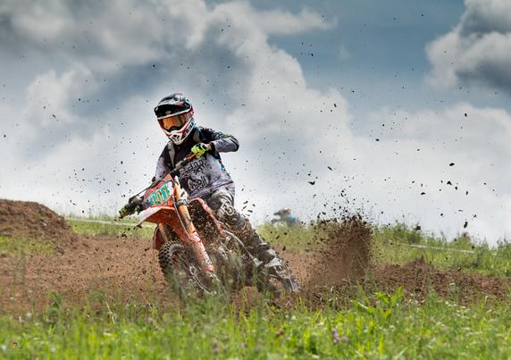 Color photo in landscape format. It shows a motocross rider on a dirt track shortly after a turn. The motorcycle is tilted slightly to the right. The rear wheel is kicking up a lot of dirt. Individual clumps of dirt are scattered up in the air across the entire image.