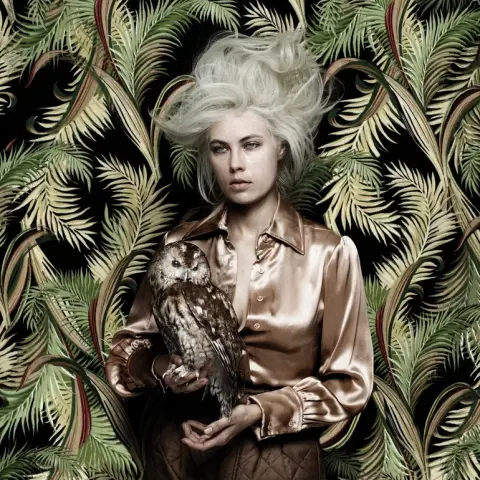 Album cover for The Day Before Halloween by Atmosphere. The artwork features a woman with platinum blonde hair teased upward, giving a wild, ghostly appearance. She wears a silky golden-brown shirt and holds a tawny owl in her hands, staring directly forward with a haunting, expressionless gaze. Behind her is a wallpaper of dark jungle-green palm fronds, blending into her form and giving the image a surreal, unsettling quality. The imagery evokes themes of eerie stillness, the uncanny, and a blend of natural and supernatural.