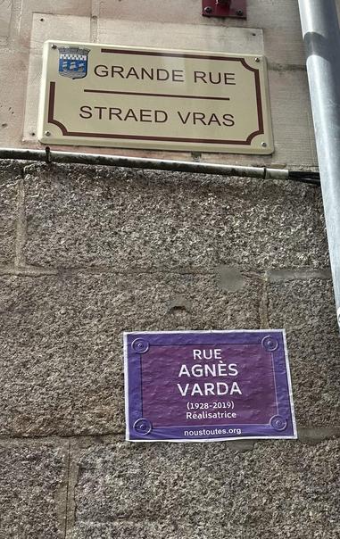 Plaque de La grande rue (strates vras) à Redon avec en dessous une affiche du collectif @noustoutes.org au fromat plaque de rue violet qui rebaptise la rue au nom d’Agnés Varda (1928-2019) réalisatrice.