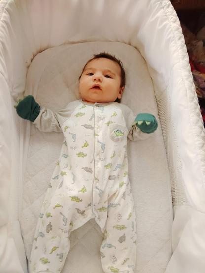 A baby with dark hair in a dinosaur print onesie with little dinosaur paw hand coverings to keep him from scratching his face. He’s in a bassinet.