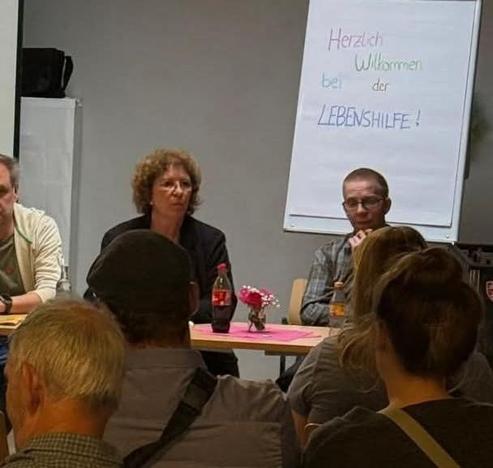 Foto aus dem Publikum heraus von der Gesprächrunde. Man sieht mehrere Hinterköpfe. Weiter vorn sitzen drei Leute an einem Tisch dem Publikum zugewandt. Rechts sitzt der Piratenkandidat Lars Wind.
Im Hintergrund steht ein Cardboard, auf dem steht: "Herzlich willkommen bei der Lebenshilfe!"