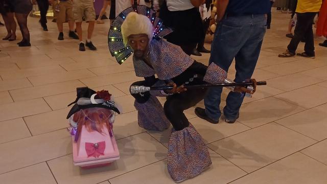 A small pink Mouse droid from Star Wars, wearing a pink and purple wig and Ironmouse's Witch of Twitch hat, standing next to someone cosplaying Ruby Rhod of The Fifth Element, who is holding a microphone toward her.  Rhod's outfit fabric uses the blue, red, and gold carpet pattern previously used in the Marriott Marquis in Atlanta (now a classic part of the convention's culture).