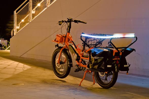 an orange Rad electric bicycle is parked facing left slightly away from the viewer. It is in front of a concrete stairway. The cargo area has a bright string of multi color LEDs that shine brightly.
