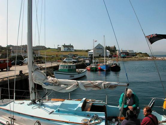 Longhope on Hoy in Orkney. The image shows a view from a harbour back over a moored yacht in the foreground which has three people sitting in the back of it. There’s open water on the right, while on the left a concrete quay extends back towards the land. It has several small boats moored to it and cars parked on it. At the landward end is a lifeboat station with other buildings in the distance. The sky is blue.