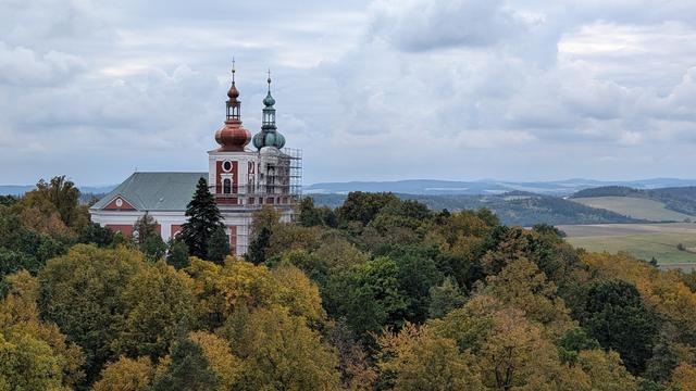 Poutní kostel Panny Marie sedmibolestné na Cvilíně z rozhledny. Nad vrcholky stromů vystupuje střecha a dvě věže. Jedna věž s nově opravenou měděnou střechou (oranžová) a druhá věž s lešením a se stále ještě zelenou oxidovavou střechou.