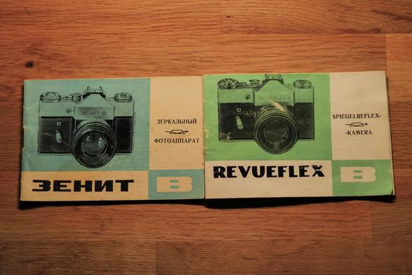 Two color-printed manuals on yellowed paper side by side: on the left, the Zenit-B manual with teal cover, Cyrillic text, and camera illustration; on the right, the Revueflex-B manual with green cover, German text ‘Spiegelreflexkamera,’ and the same camera illustration under the West German export name Revueflex. (Photo by me)