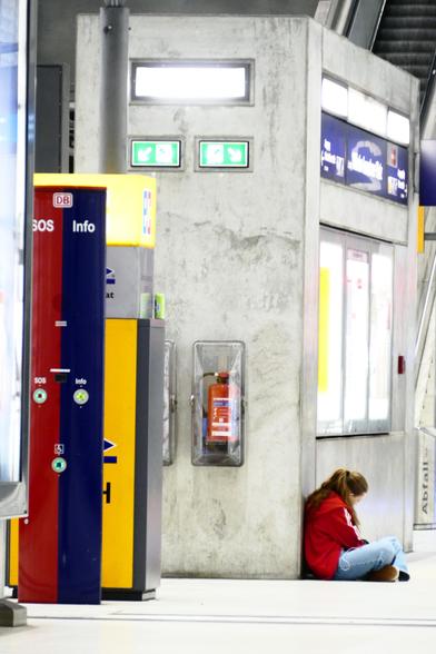 Schräg von der Seite ist ein Mädchen mit roter Jacke abgelichtet, das auf einem deutschen hellgrauen Bahnsteig im Schneidersitz an einer Betonwand lehnt und etwas liest, während es augenscheinlich Musik hört. Über ihm sind in ein dunkelblauer Wegweiser sowie ein hell leuchtender Fahrplan-Schaukasten in die Wand eingelassen, und links neben dem Mädchen hängt ein feuerroter Feuerlöscher an der Wand. Daneben wiederum steht ein dunkelgelber Geldautomat, der von einer schmalen, hälftig dunkelroten und hälftig dunkelblauen SOS- bzw. Infosäule verdeckt wird. Die pastelligen Farben stellen einen ästhetischen Kontrast zur betonhellgrauen Umgebung dar. Da die große Brennweite die Szenerie ihrer räumlichen Tiefe beraubt hat, wirkt das Foto eher wie ein modernes Gemälde.