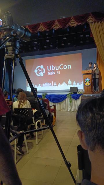 UbuCon main hall stage, with 2 hosts standing at the podium (on the right side of the screen), and attendees sitting in chairs.