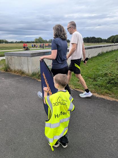 A child wearing a bright yellow vest with reflective stripes, labeled "Helfer" and "parkrun" on the back, is holding a sign. The child is walking alongside two adults on a paved path in a park. The adults are casually dressed, and the scene is set on a cloudy day with other people visible in the background, some walking and others sitting on a wall. The surroundings include grassy areas and a large open field.