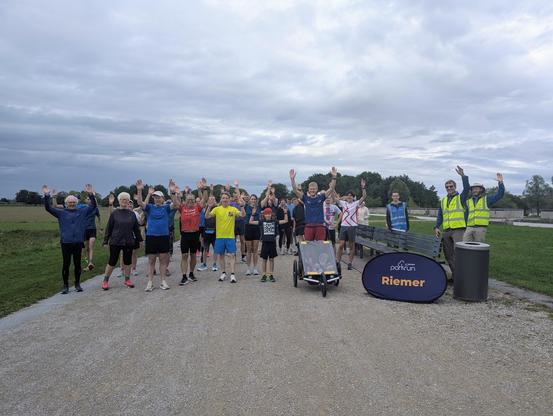 Group of people standing on a gravel path in a park, waving with their hands. Some are wearing athletic clothes, and two individuals on the right are wearing high-visibility vests. There is a sign with "parkrun Riemer" on the ground.