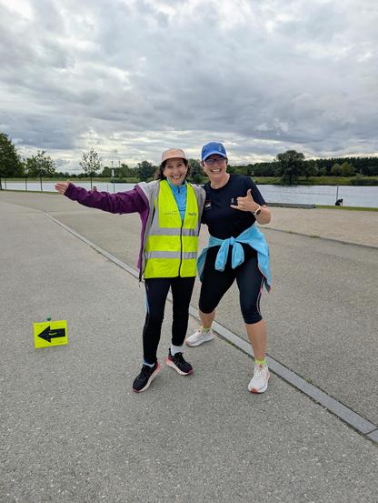 Two women standing on a paved area near a body of water. One woman is wearing a high-visibility vest and a pink cap, pointing to the left. The other woman is wearing a black shirt, black cap, and a blue headscarf, giving a thumbs-up. Both are smiling and appear to be dressed for running or walking. There is a small yellow sign with a black arrow pointing to the left on the ground in front of them. The sky is cloudy, and trees are visible in the background.